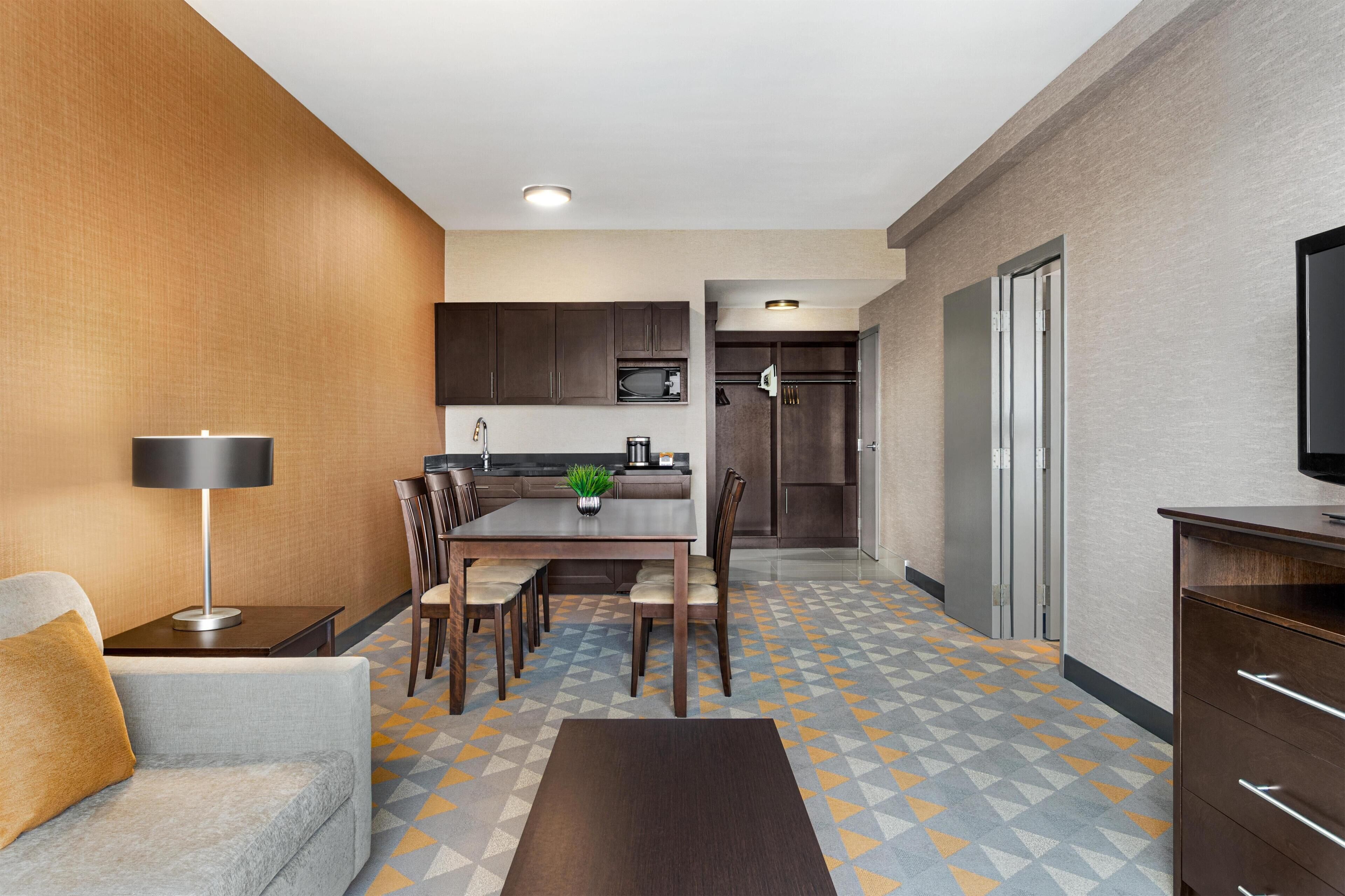 A modern hotel room with a dining area, kitchenette, and a TV on the right.