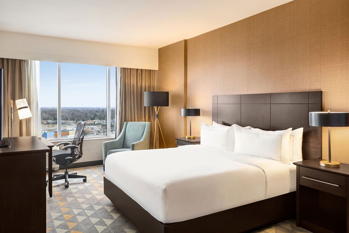 Habitación de hotel con una cama grande, dos mesitas de noche con lámparas, un escritorio con una silla y vistas a la ciudad a través de amplios ventanales.