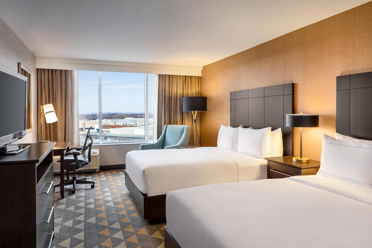 Ein Hotelzimmer mit zwei Betten, einem Schreibtisch, einem Stuhl, einem Fernseher und einem großen Fenster mit Blick.