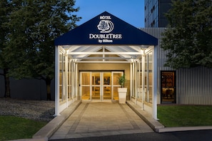 L’entrée d’un hôtel avec une façade en verre, une enseigne au-dessus indiquant « HÔTEL DOUBLETREE by Hilton », et un chemin bien éclairé menant à l’entrée.