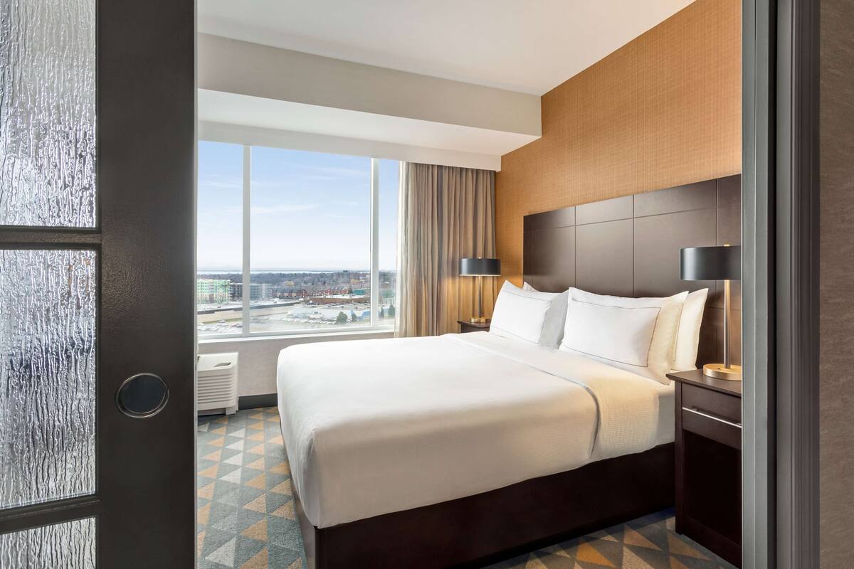 Ein Hotelzimmer mit einem großen Bett, einem Nachttisch, einer Lampe, einem Fenster mit Blick auf die Stadt und einer Tür.
