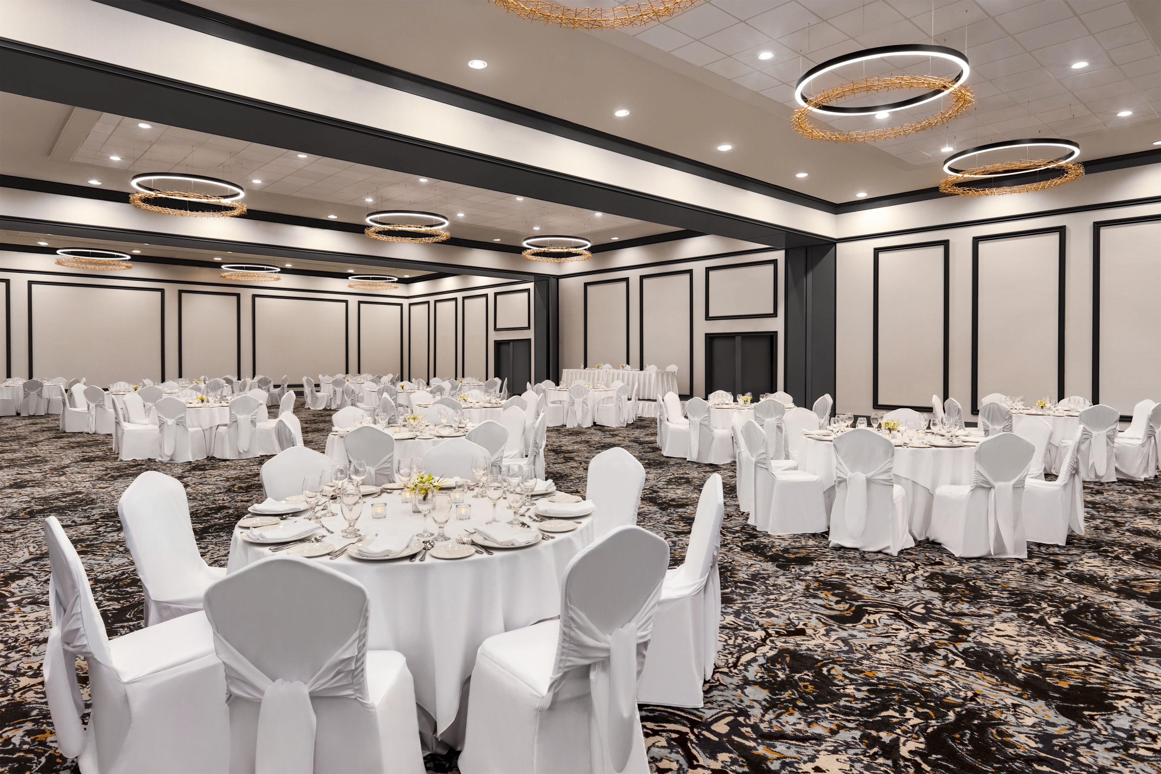A large, well-lit banquet hall with round tables, white chairs, and modern chandeliers.