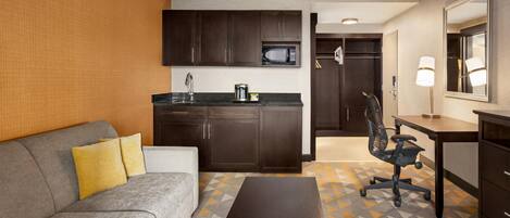 A compact hotel room with a kitchenette, a sofa, a desk, and a chair.