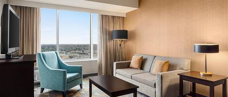 A hotel room with a sofa, armchair, coffee table, TV, and a view of the cityscape.