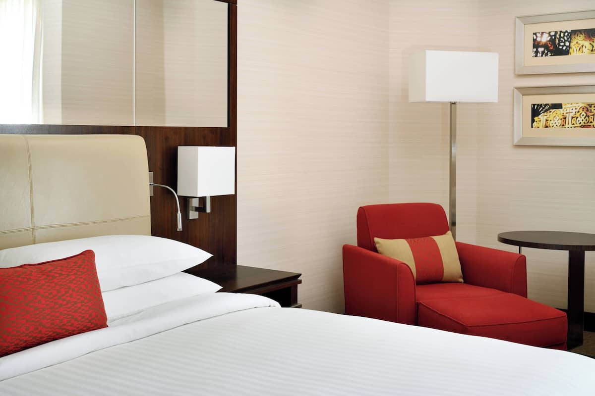 Habitación de hotel con dos camas, un sillón rojo, una mesita pequeña y una lámpara de pared.