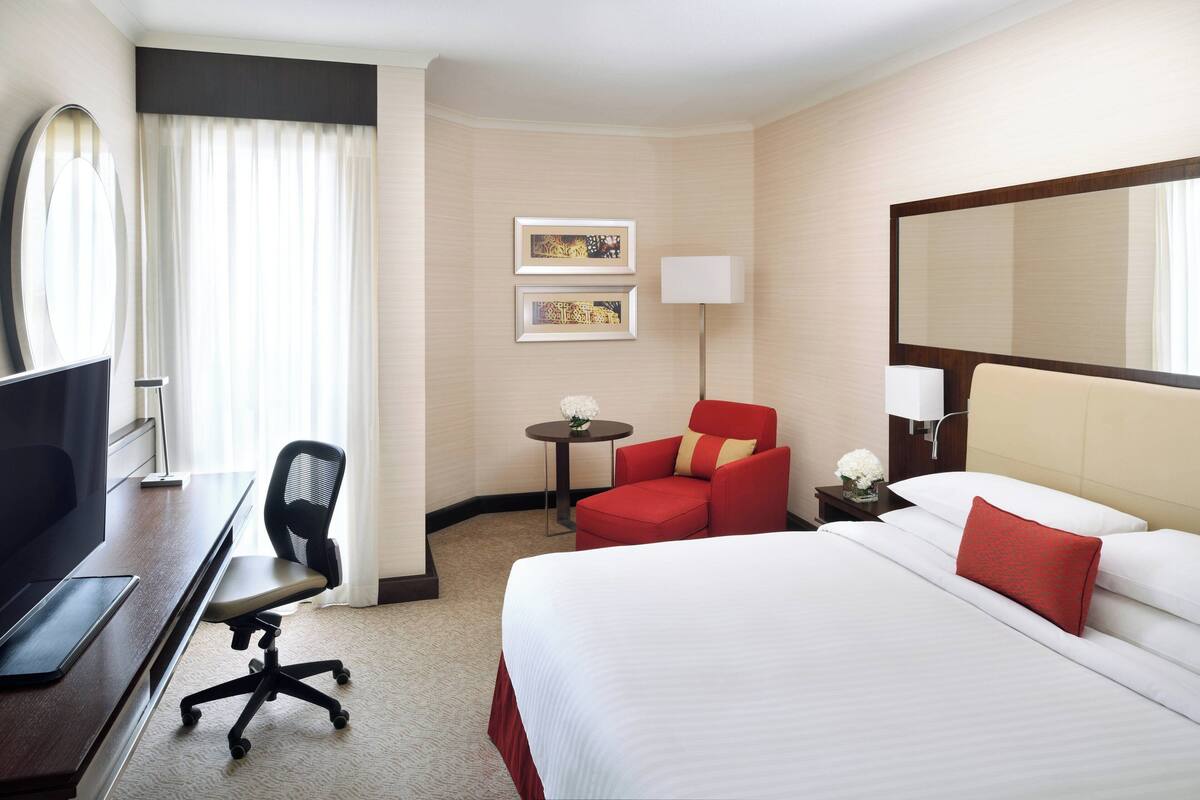Habitación de hotel con una cama grande, un escritorio con una computadora, una silla roja, una mesita con una lámpara y dos cuadros en la pared.