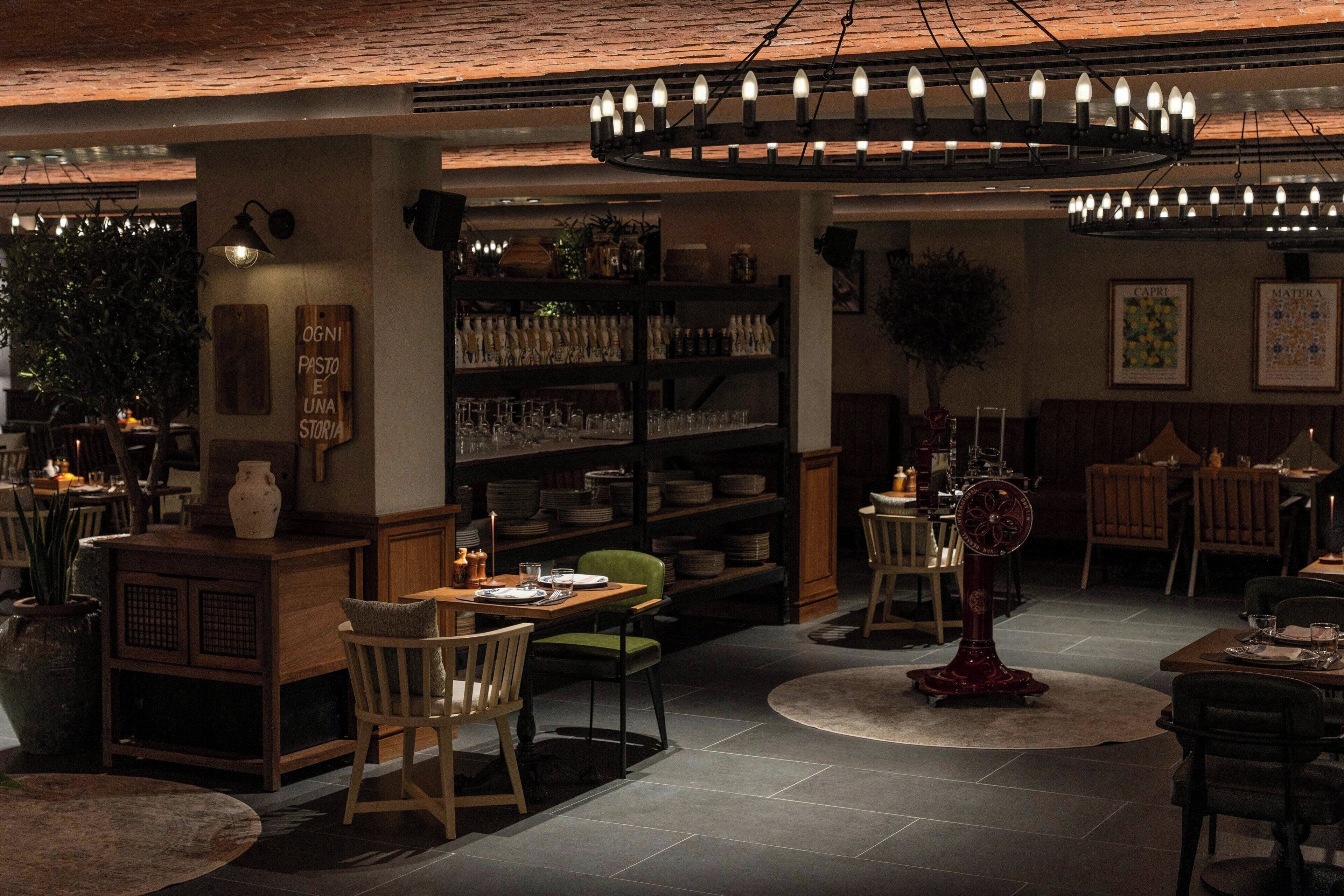 A restaurant interior with wooden furniture, a central chandelier, and a bookshelf filled with plates and glasses.