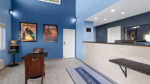 A reception area with a blue wall, two posters, a reception desk, a wooden chair, a bench, and a doormat with "Travelodge" branding. - Travelodge by Wyndham Banning CA Near Casino/Outlet Mall (Banning)