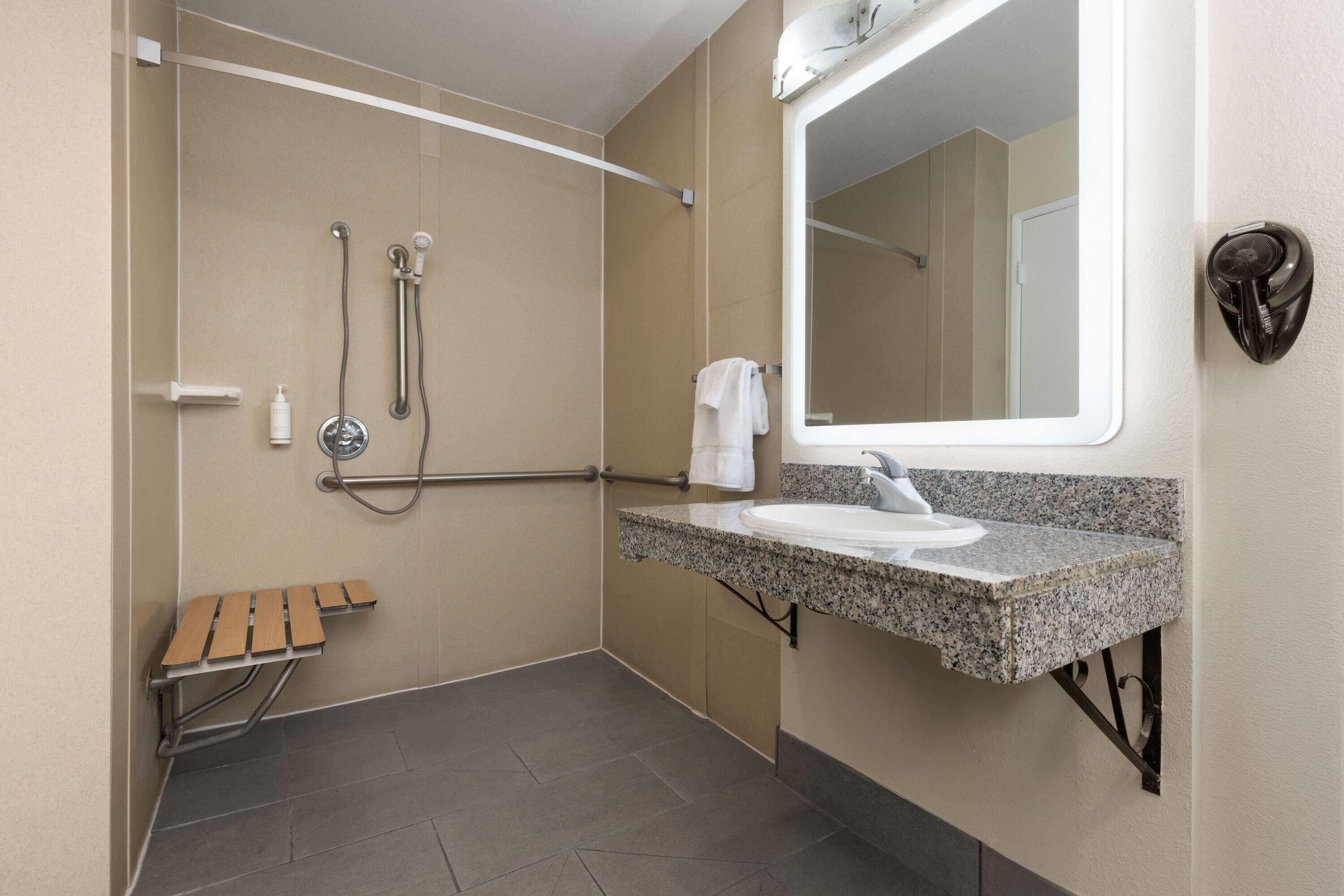 A modern bathroom with a shower, grab bar, bench, sink, and mirror.