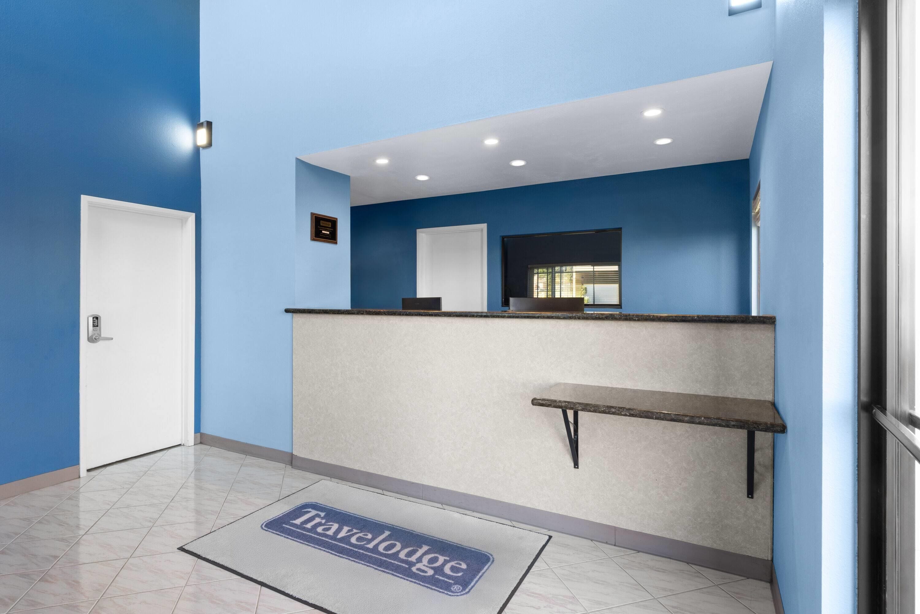 A reception area with a blue wall, a reception desk, a white door, and a 'Travelodge' mat on the floor.