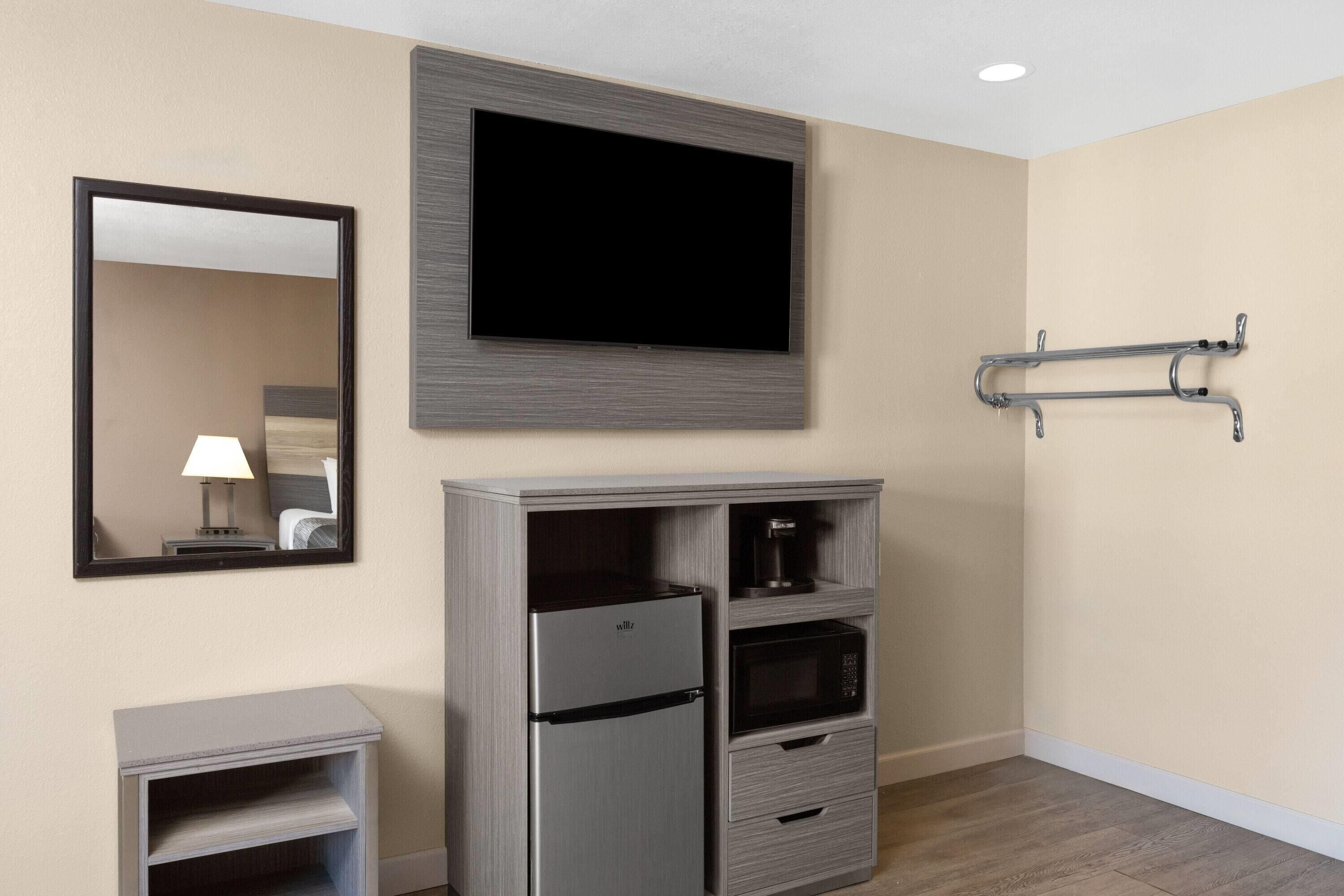 A hotel room with a flat-screen TV mounted on the wall, a minibar, a refrigerator, a microwave, a small table with a lamp, and a coat rack.