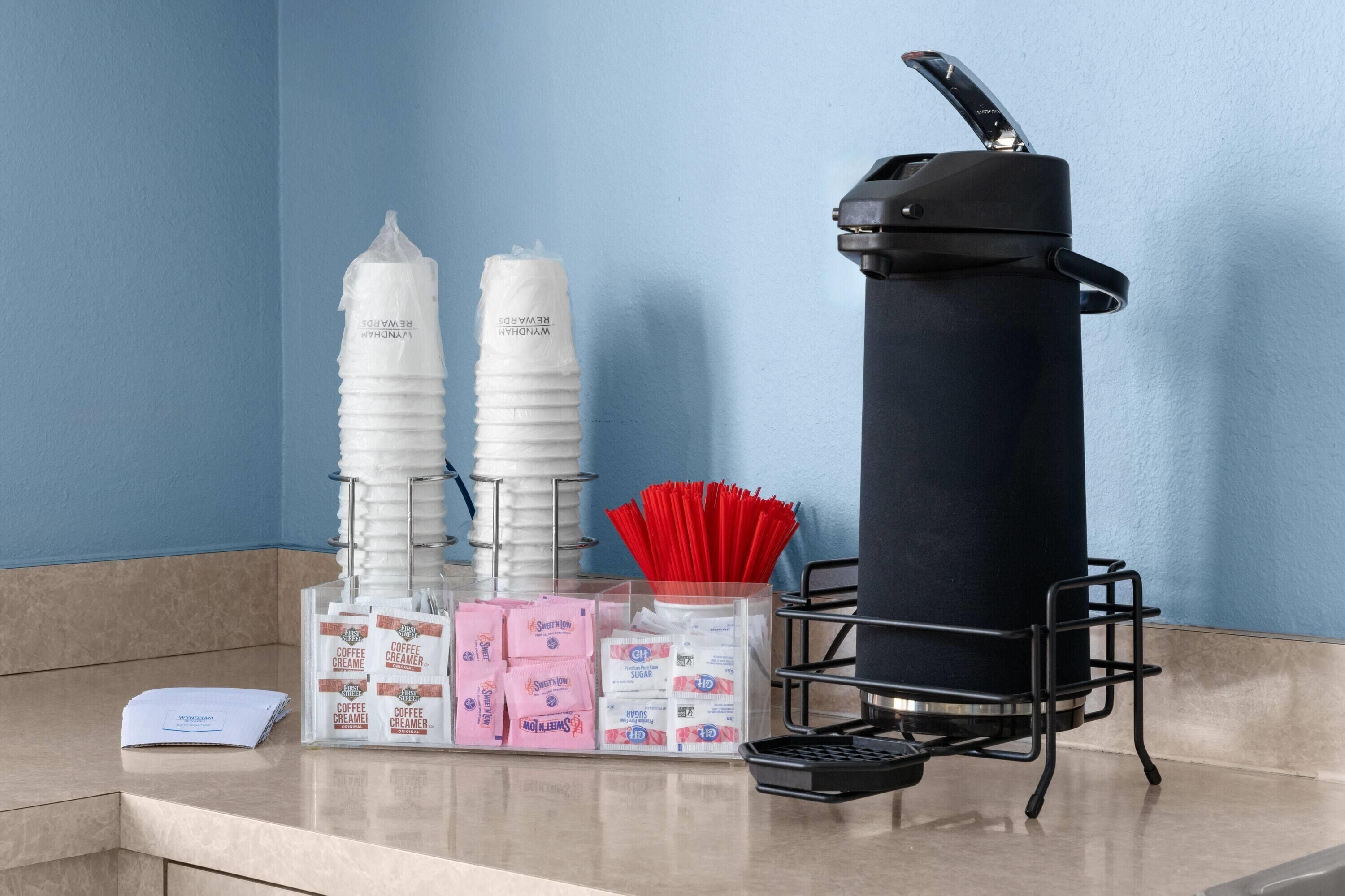 A coffee station with disposable cups, a coffee creamer dispenser, sugar packets, and a trash bin.