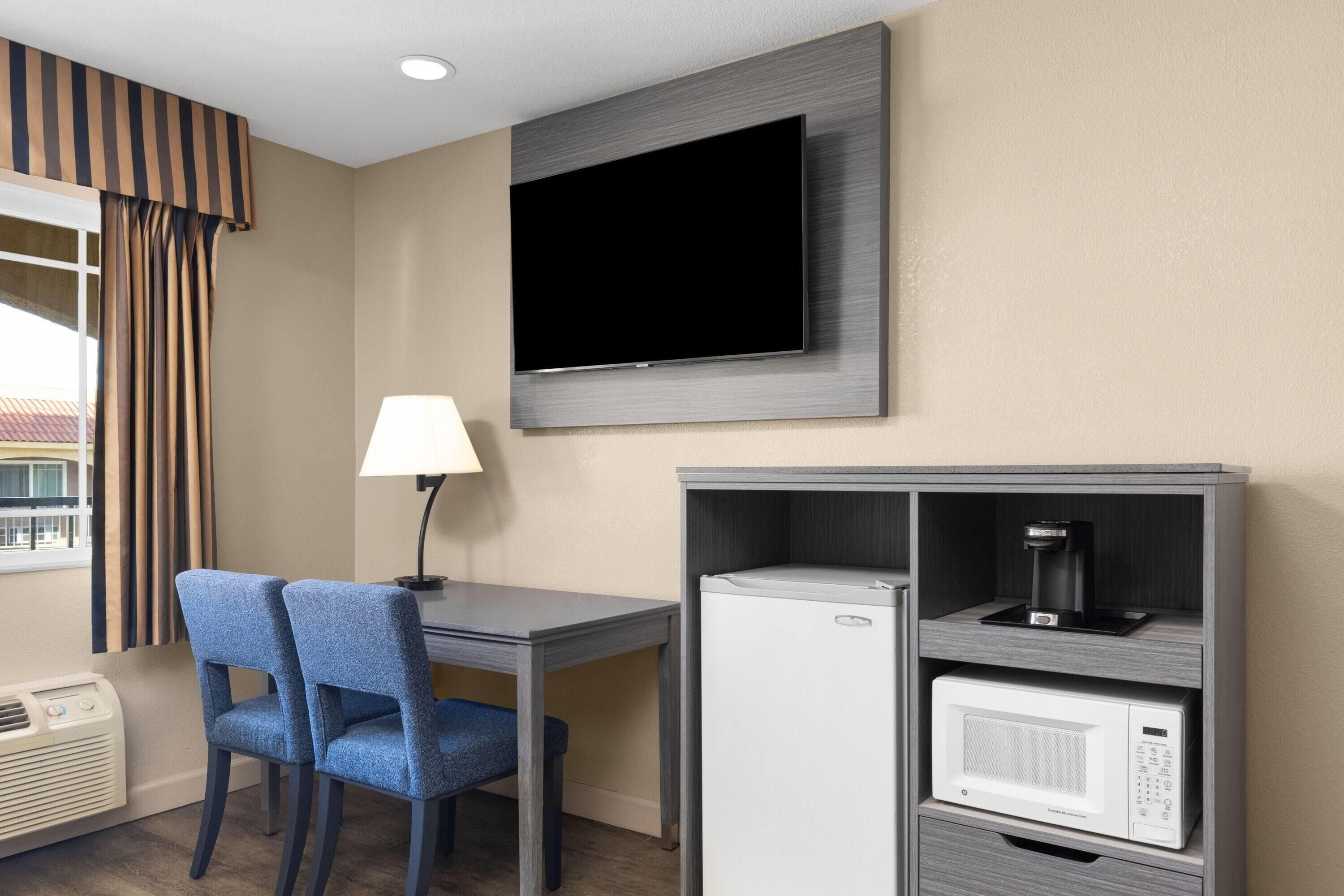 A hotel room with a flat-screen TV mounted on the wall, a desk with two blue chairs, a lamp, a mini-fridge, and a microwave.