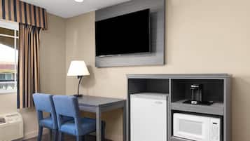 A hotel room with a flat-screen TV mounted on the wall, a desk with two blue chairs, a lamp, a mini-fridge, and a microwave.