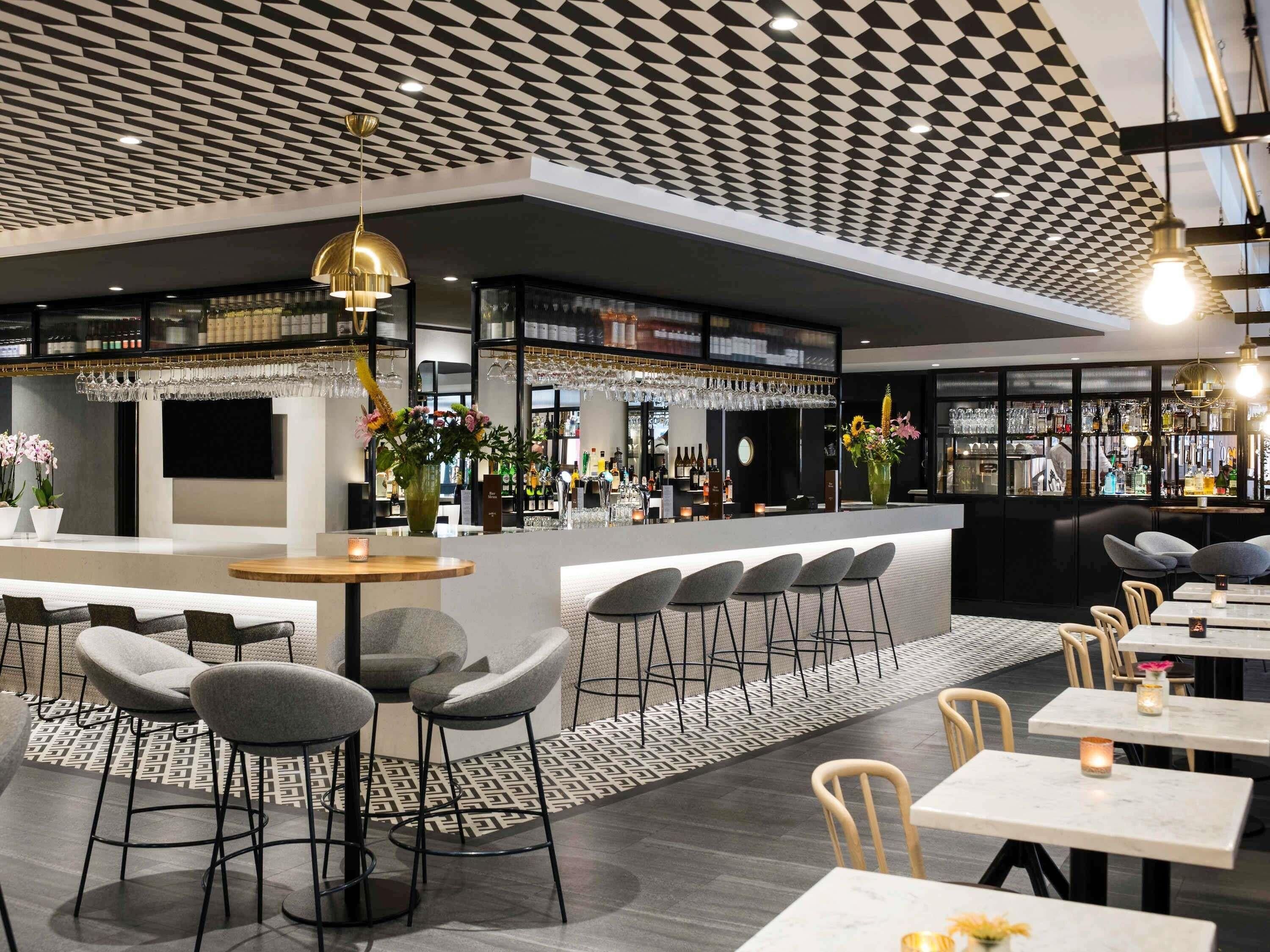 A modern bar area with a geometric ceiling, bar stools, and a seating area with tables and chairs.