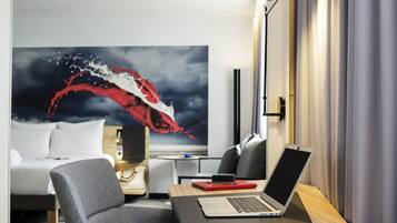 A modern hotel room with a laptop on a desk, a large abstract painting on the wall, and a bed with a striped rug.