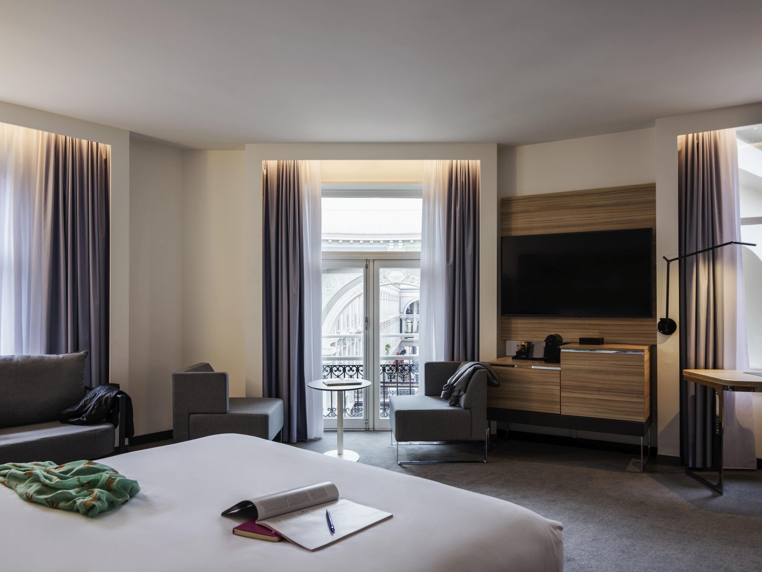 A modern hotel room with a large bed, a desk, a TV, and a balcony with a view of the city.