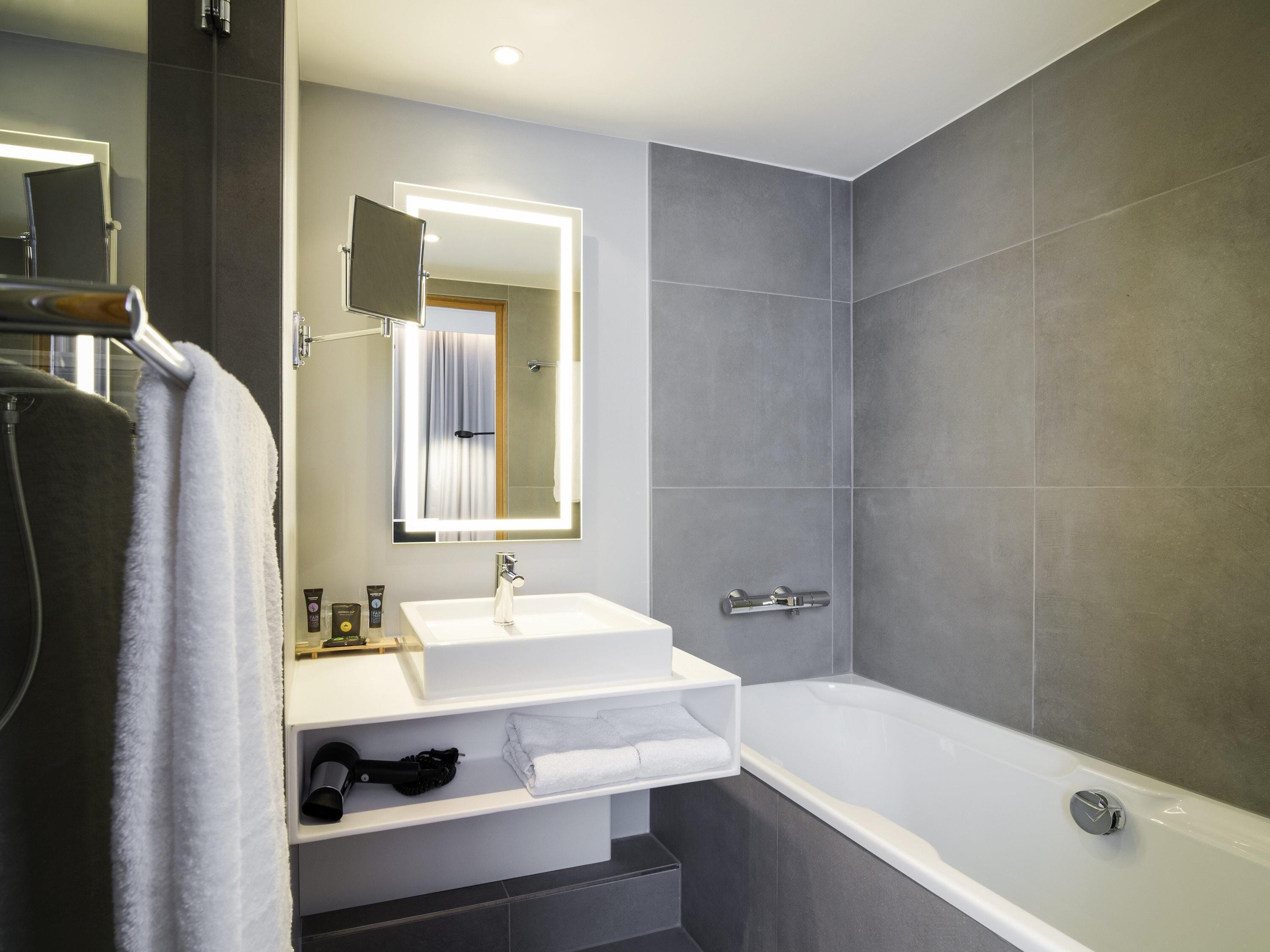A modern bathroom with a white sink, a mirror, a bathtub, and grey tiles.