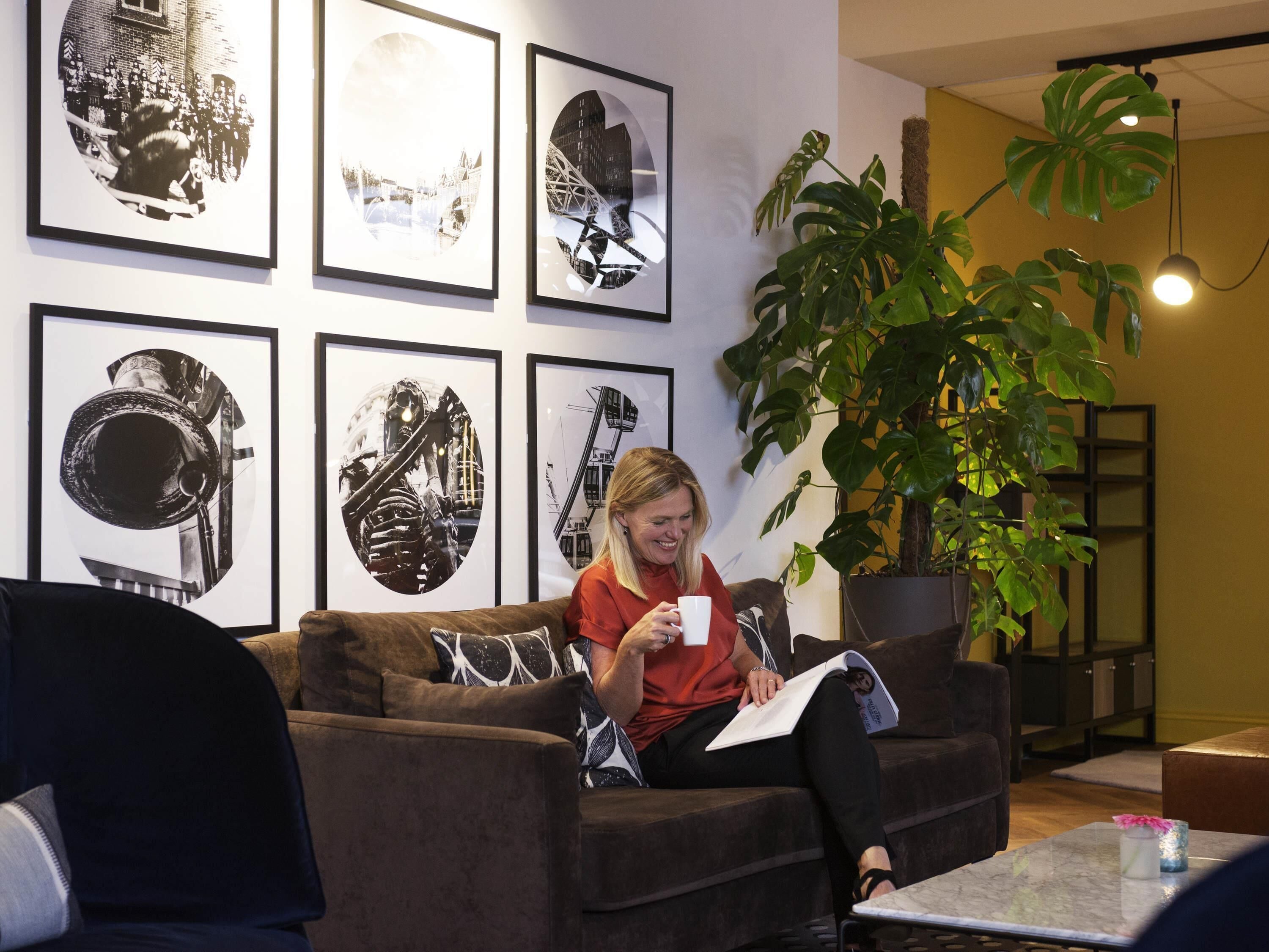 A person sitting on a brown couch in a room with framed pictures on the wall, a large potted plant, and a small table with a cup and a book.