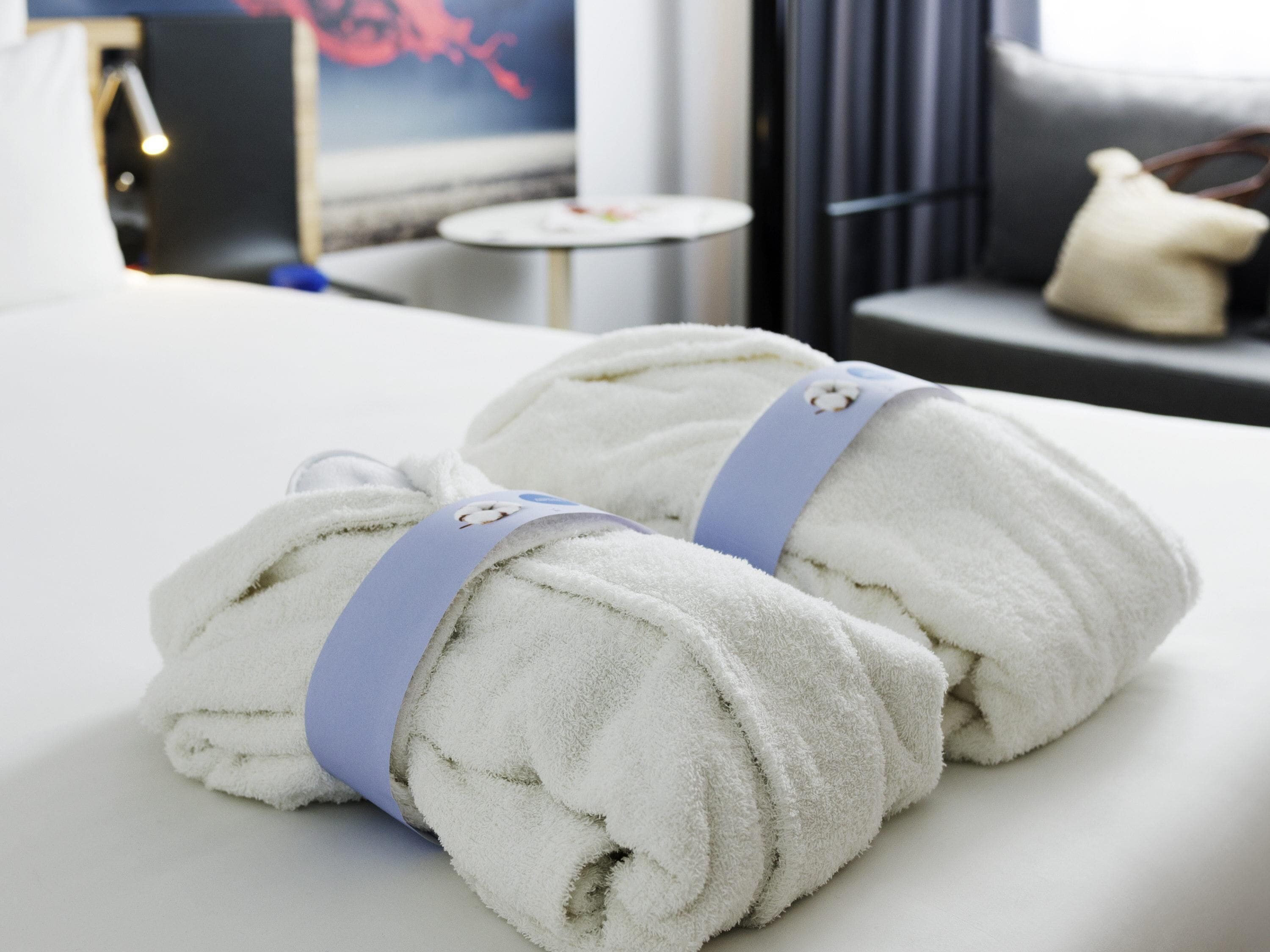 Una cama bien hecha con dos toallas enrolladas sujetas con un lazo azul, una mesita redonda con una lámpara y un sofá gris con un cojín decorativo.