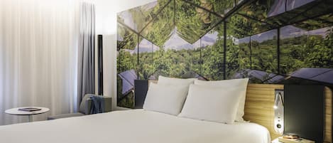 Habitación de hotel con una cama grande, una mesita de noche, una lámpara y vistas a un bosque a través de un diseño de ventana geométrico.