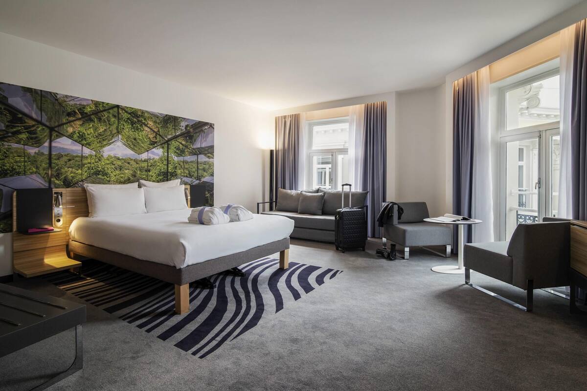 Una habitación de hotel moderna con una cama grande, un sofá, un escritorio y vistas de un paisaje urbano a través de la ventana.