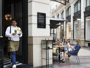 A waiter carrying a tray with a drink and a glass of water, a sign with text, and a building facade with columns and windows. - Novotel Den Haag City Centre (The Hague)