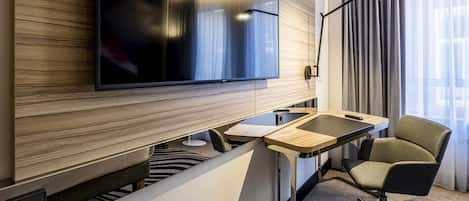 Una habitación de hotel moderna con un televisor de pantalla plana montado en una pared de paneles de madera, un escritorio con una silla y un ventanal con cortinas.