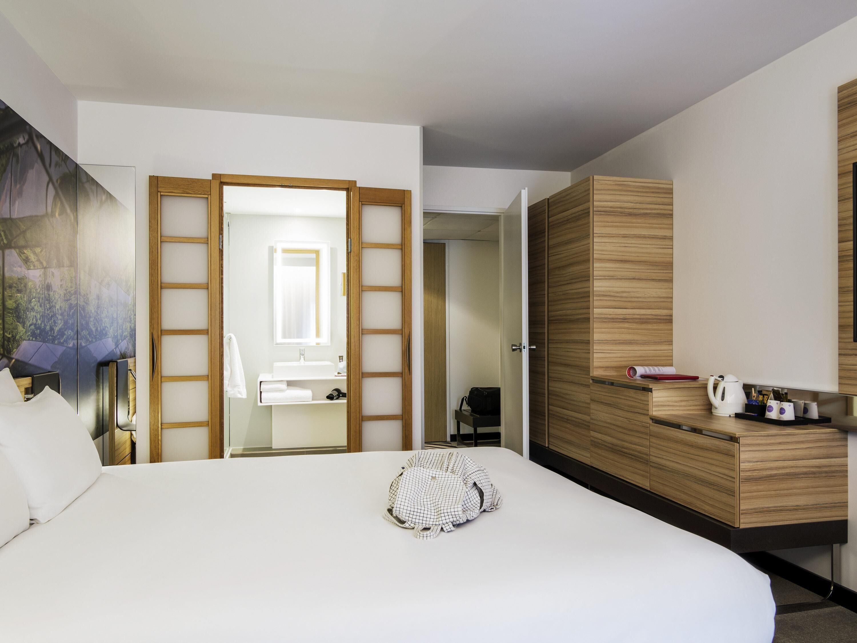 A hotel room with a bed, a wooden wardrobe, a mirror, a bathroom with a sink and towel rack, and a painting on the wall.
