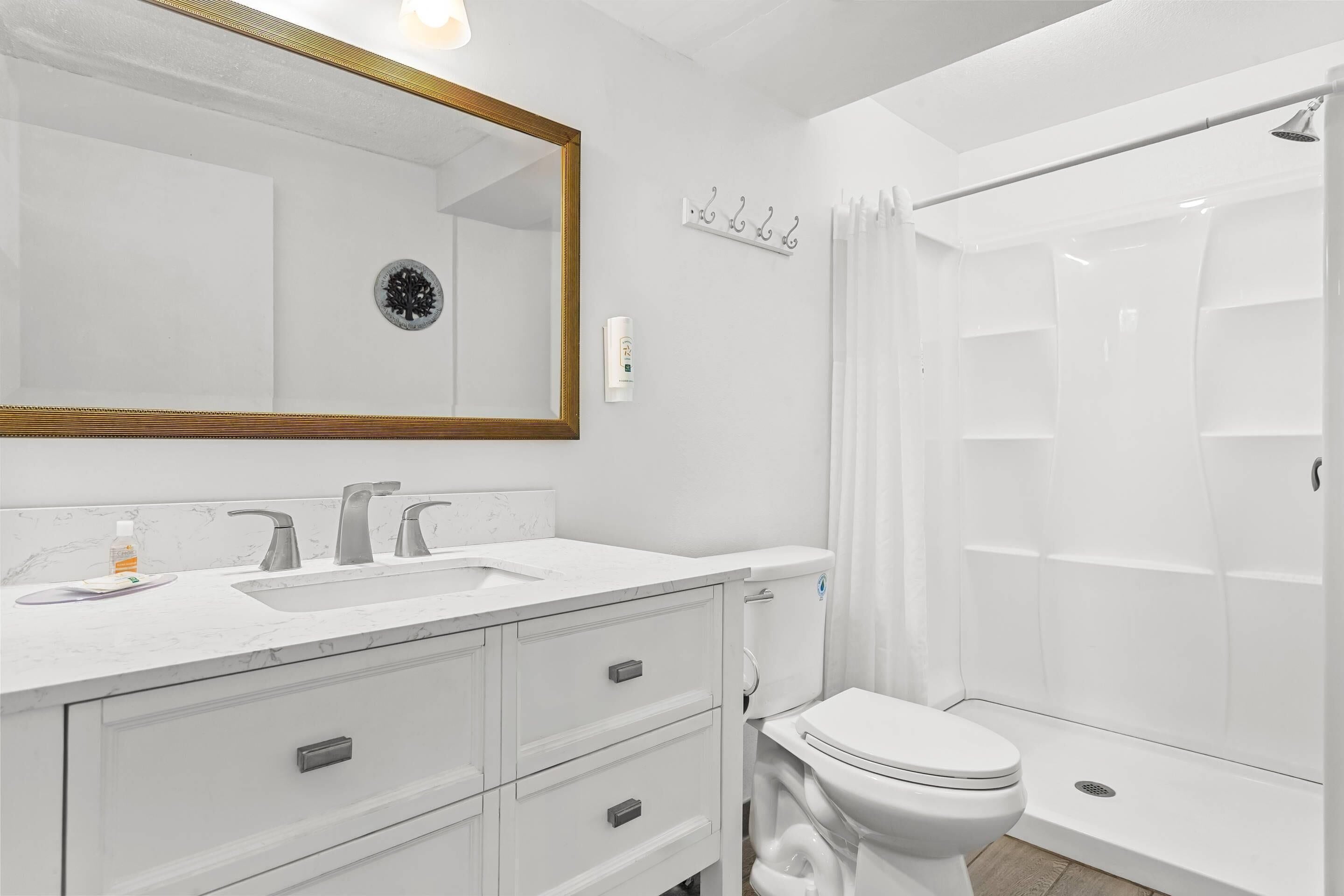 Een moderne badkamer met een wit wastafelmeubel, twee kranen, een spiegel met gouden lijst, een toilet en een douchegedeelte.
