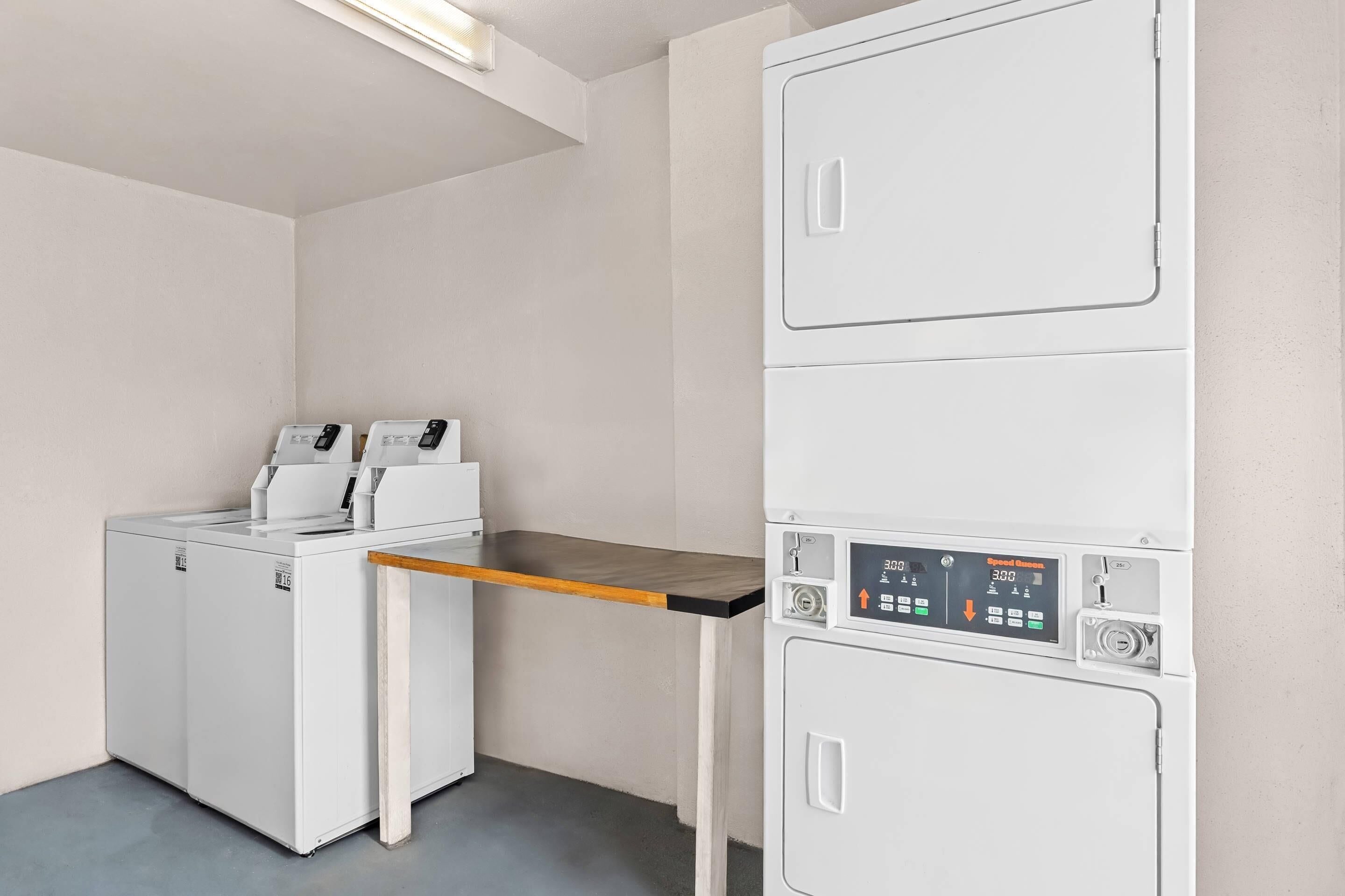 A laundry room with stacked washers and dryers, a wooden shelf, and a control panel.
