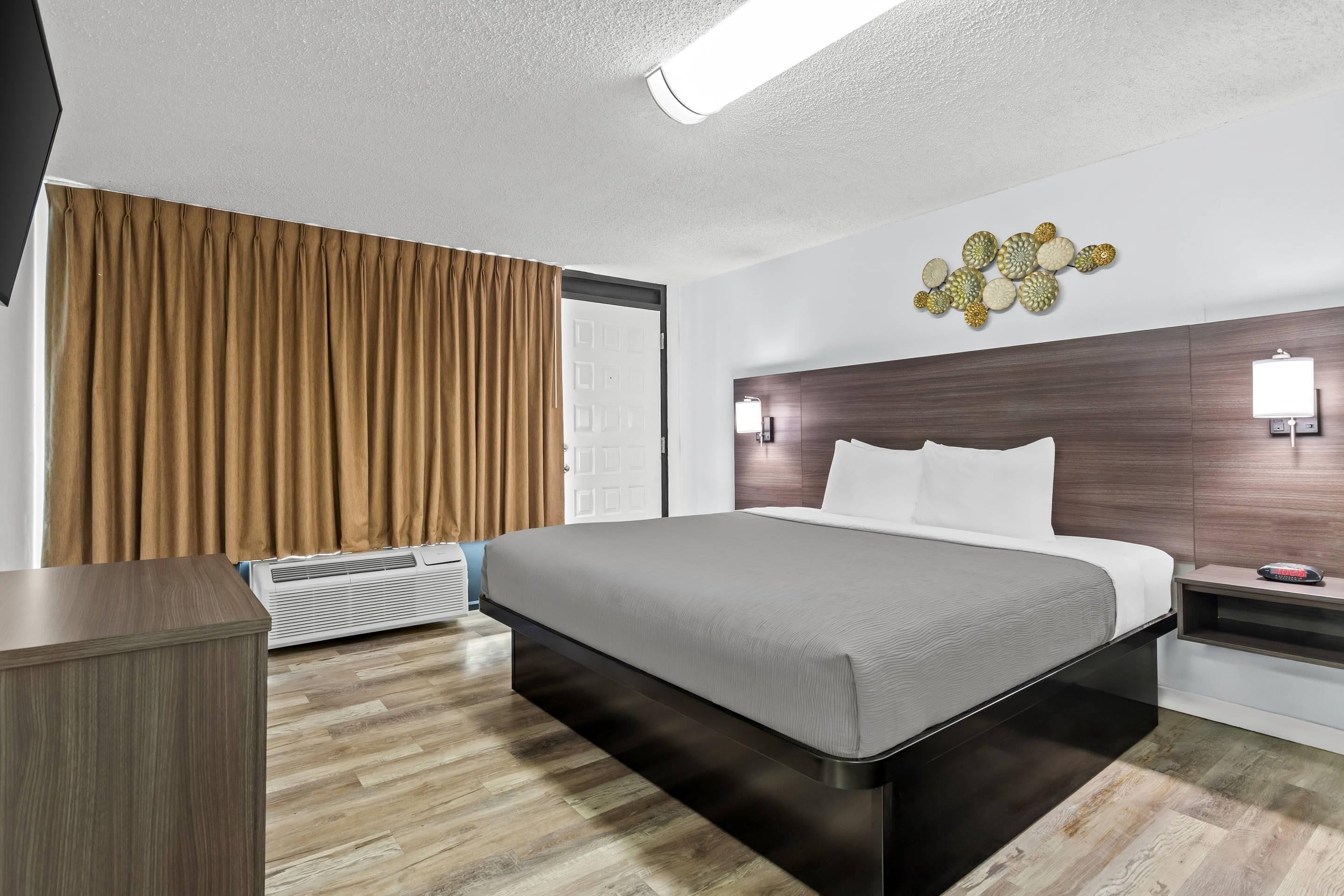 Een moderne hotelkamer met een groot bed, houten vloeren, een plafondlamp, een wandairconditioner en een decoratief kunstwerk aan de muur.