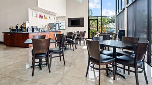 Un comedor moderno con una cocina al fondo, ventanas amplias y un televisor de pantalla plana montado en la pared.