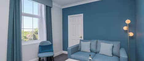 A room with a blue wall, a white door, a blue sofa with cushions, a grey chair, and a marble coffee table with a bottle and glasses.