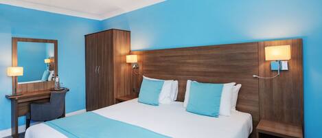 A hotel room with a large bed, a wooden wardrobe, a desk with a chair, and two wall-mounted lamps.