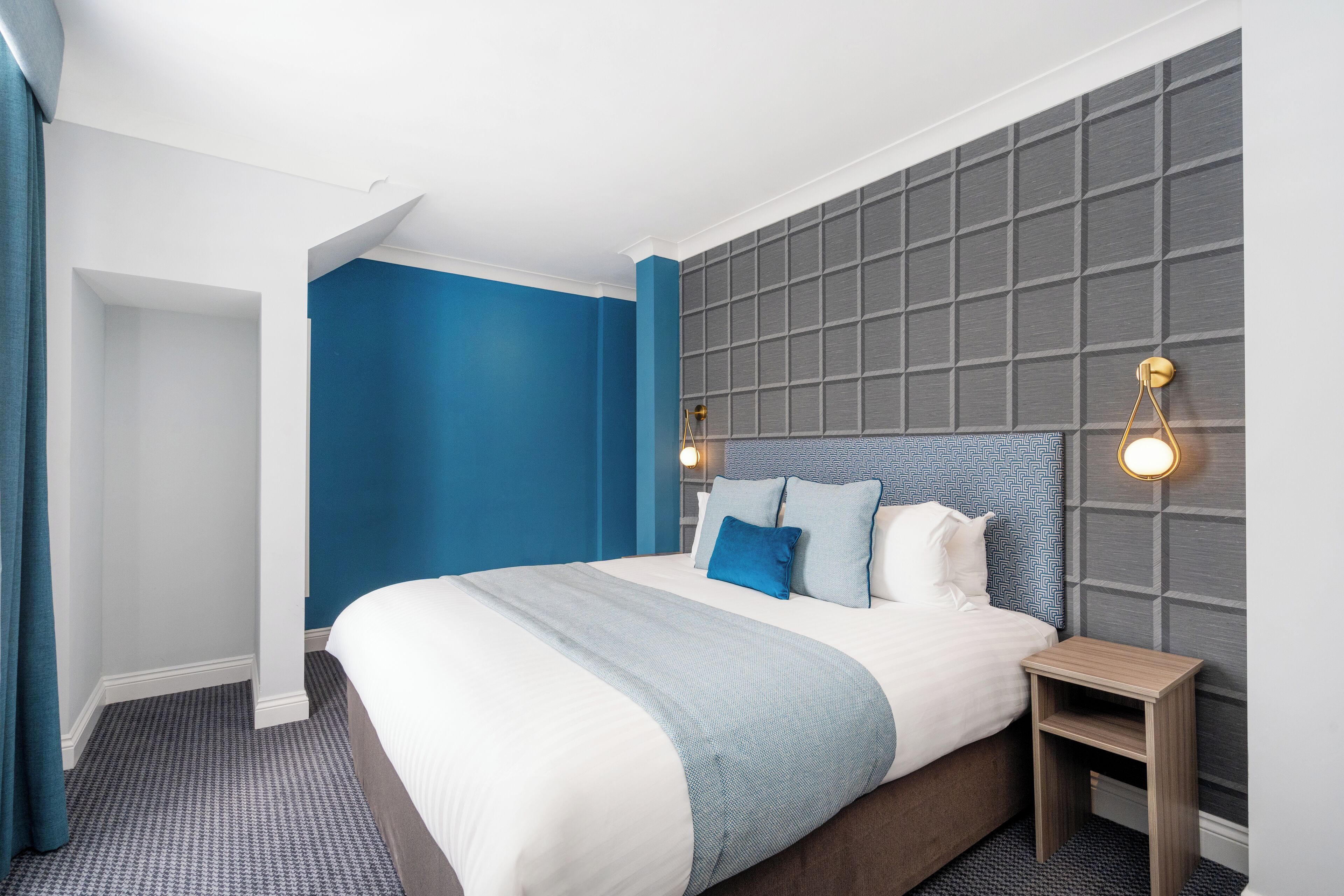 A modern hotel room with a large bed, a nightstand, and a wall-mounted light fixture.