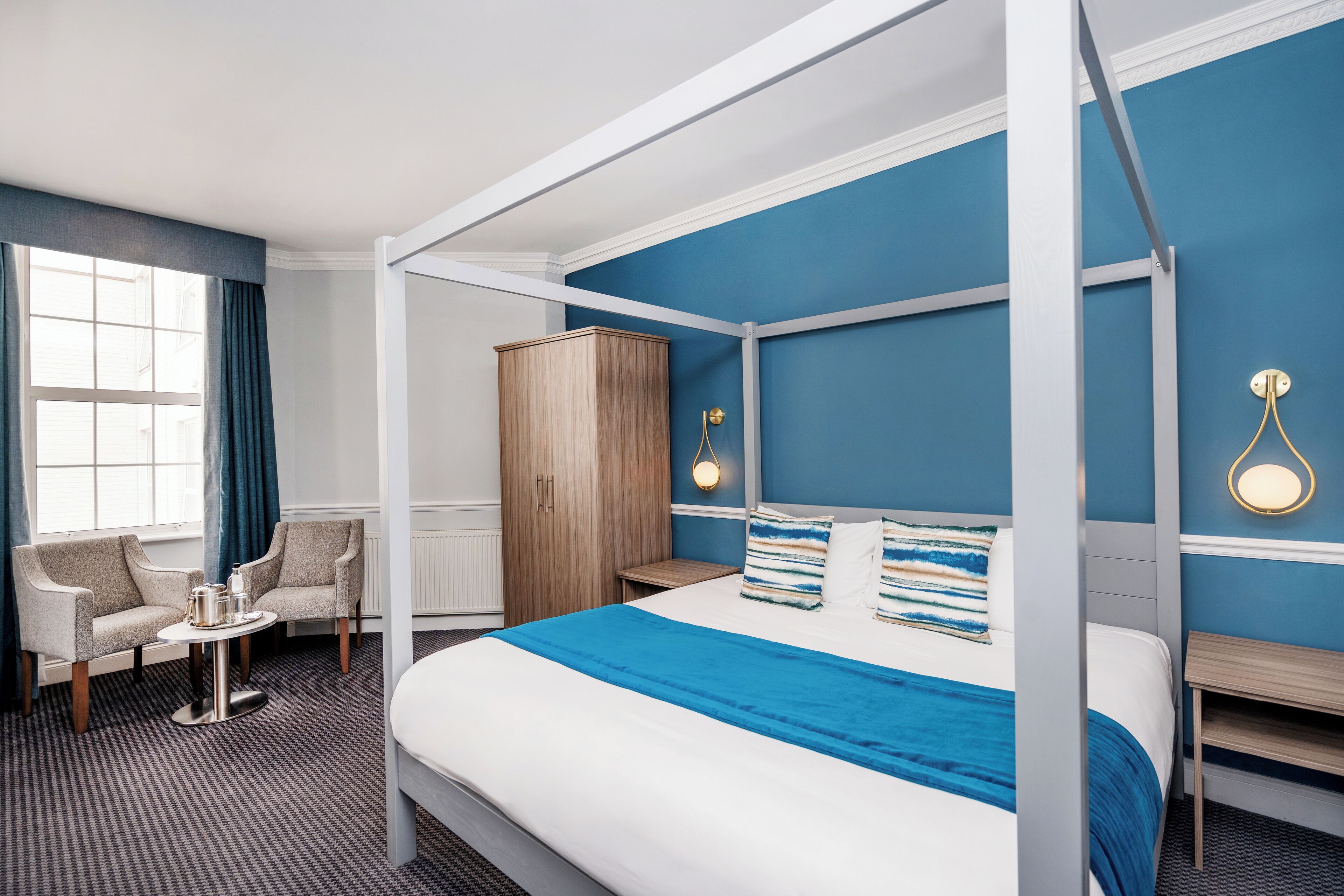Habitación de hotel con cama con dosel, zona de estar con sofá y mesita, armario de madera y ventana con cortinas.
