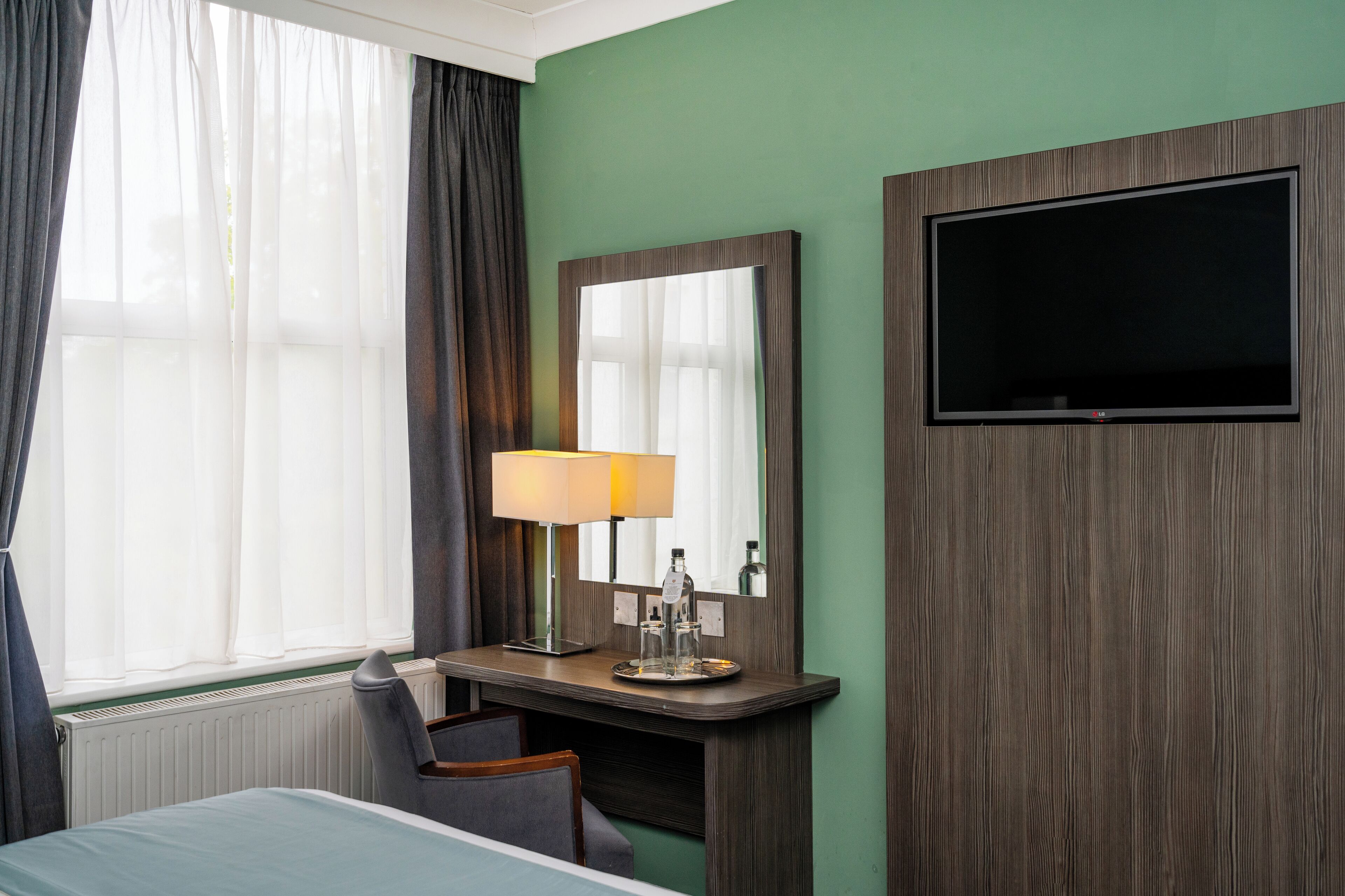 Ett hotellrum med en säng, ett skrivbord med en lampa, en tv och ett fönster med gardiner.