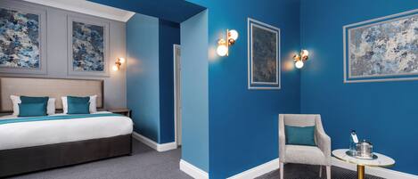 Ett hotellrum med en stor säng, en stol, ett litet bord och konstverk på väggarna.