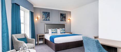 A hotel room with a bed, a desk, a chair, a nightstand, and a window with curtains.
