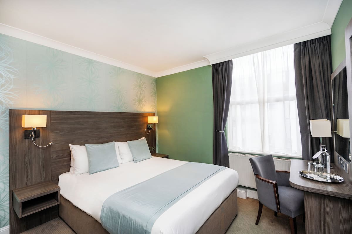 A hotel room with a large bed, bedside lamps, a desk with a tray, and a window with curtains.