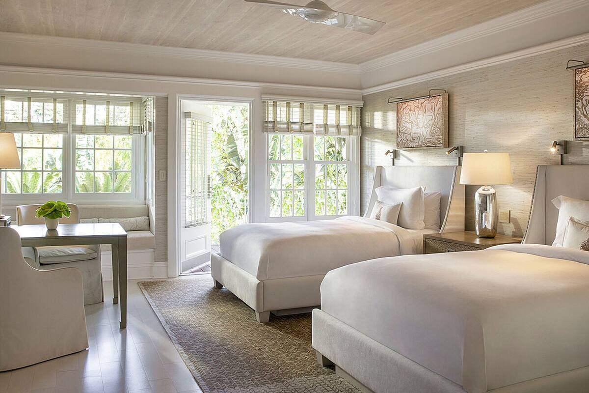 A hotel room with two beds, a desk, and a chair. There are large windows with blinds, a ceiling fan, and a rug on the floor.