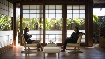 Two guests sit in a tranquil space with a view of lush tropical plants through large glass doors.