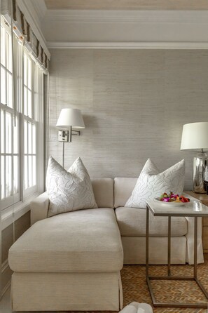 A modern living room with a beige sectional sofa, two patterned throw pillows, a small side table with a lamp, and a window with blinds.