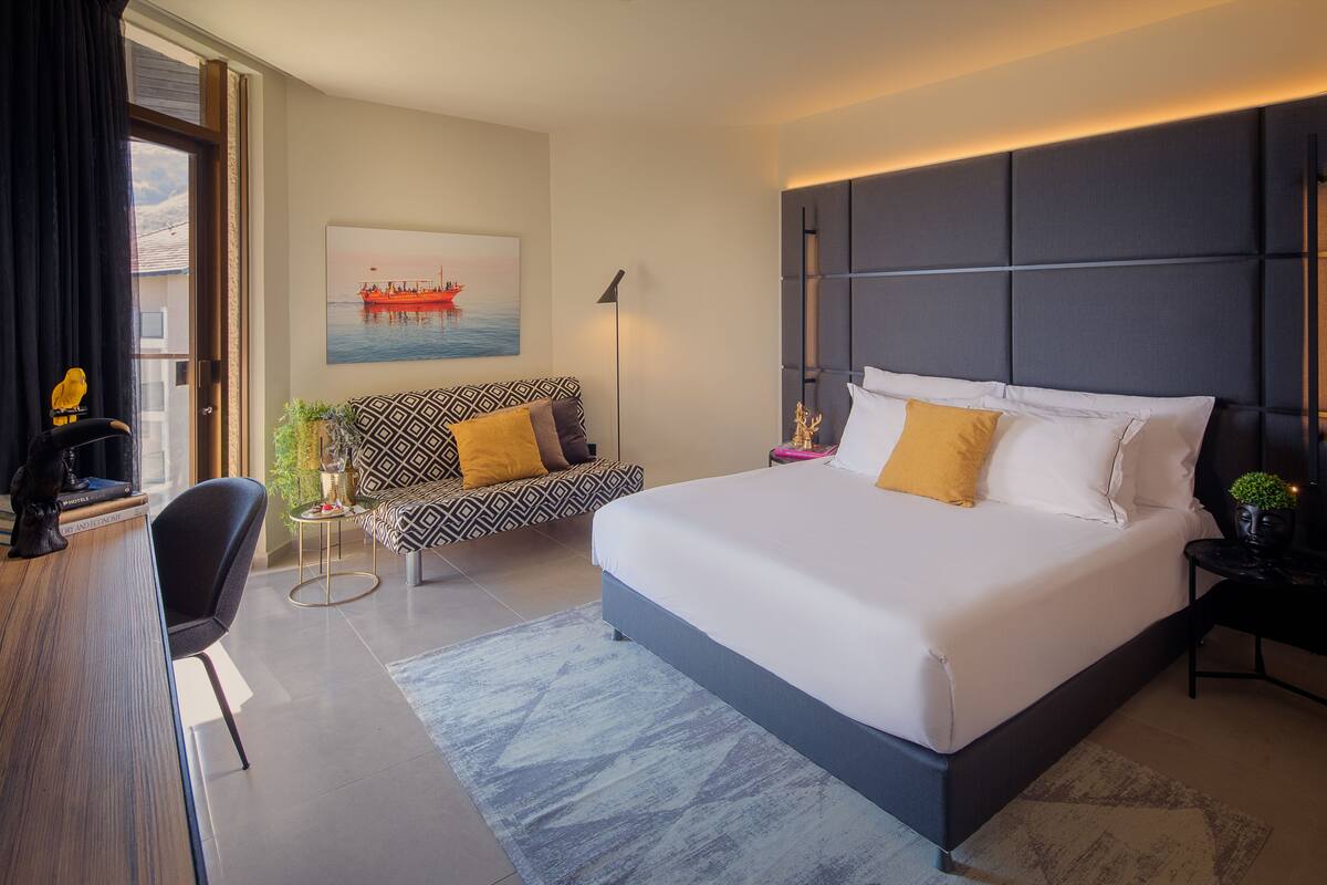 A modern hotel room with a large bed, a sofa, a desk, and a window with curtains.