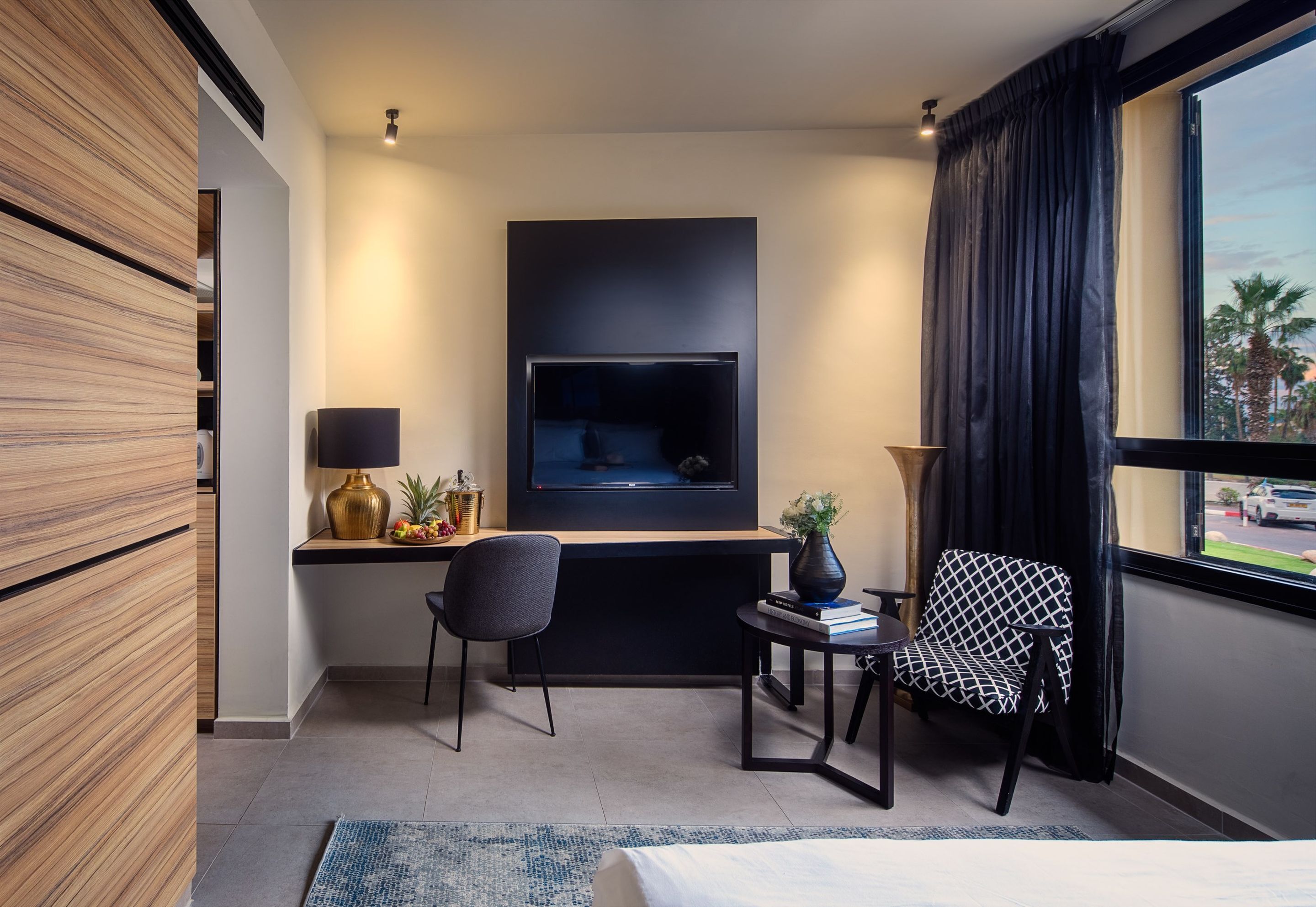 Una habitación de hotel moderna con una televisión de pantalla plana, un escritorio con lámpara, una silla, una mesita con un florero y un ventanal grande con cortinas.