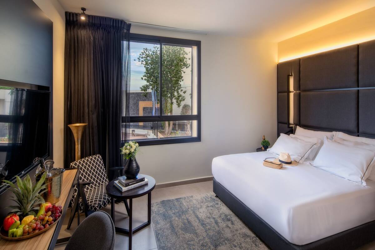 A modern hotel room with a large bed, a desk with a laptop, a small table with a vase of flowers, and a view of the outdoors through a large window.