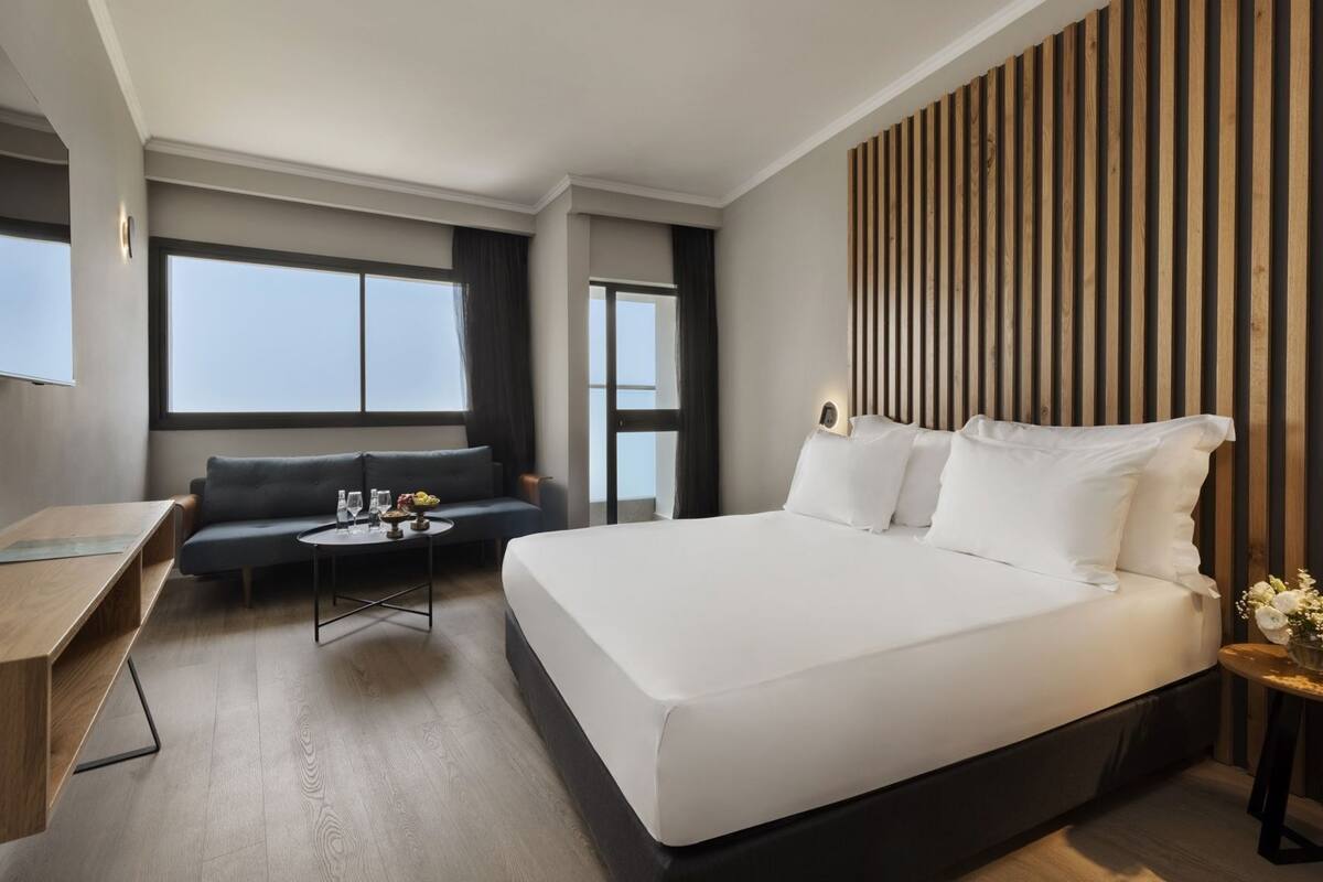 A hotel room with a large bed, a sofa, a small table with drinks, and a wooden accent wall.