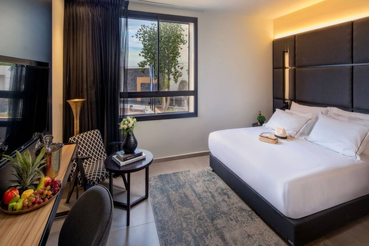 A modern hotel room with a large bed, a desk with a computer, a small side table, and a window with a view of the outdoors.