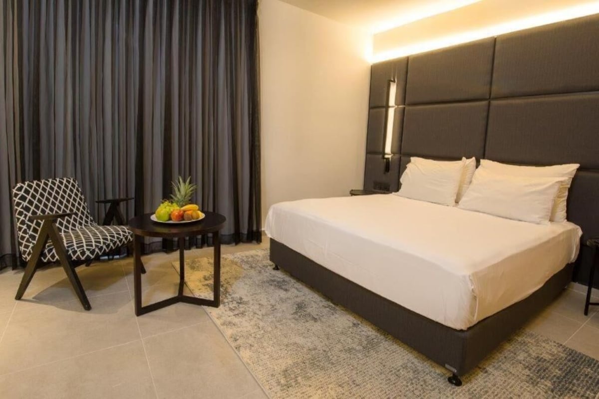 Habitación de hotel con una cama grande, una silla, una mesita con frutas y una cortina.